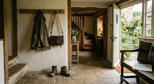 A weathered field jacket, leather satchel, and worn boots hanging in the entry hall of a historic countryside cottage, illuminated by warm natural light.