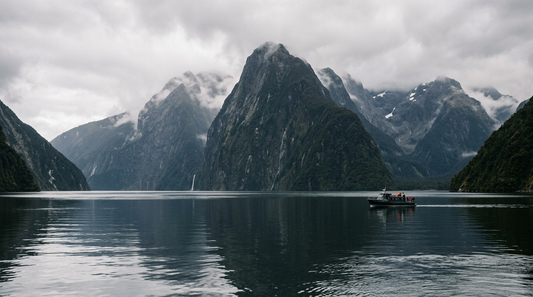 Milford Sound in New Zealand with steep mountains, low clouds, calm water, and a small boat crossing the fjord.