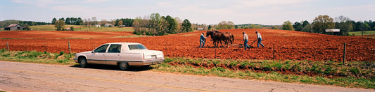 Farmers plowing a red Georgia field with draft horses beside a white Cadillac parked on a rural road.